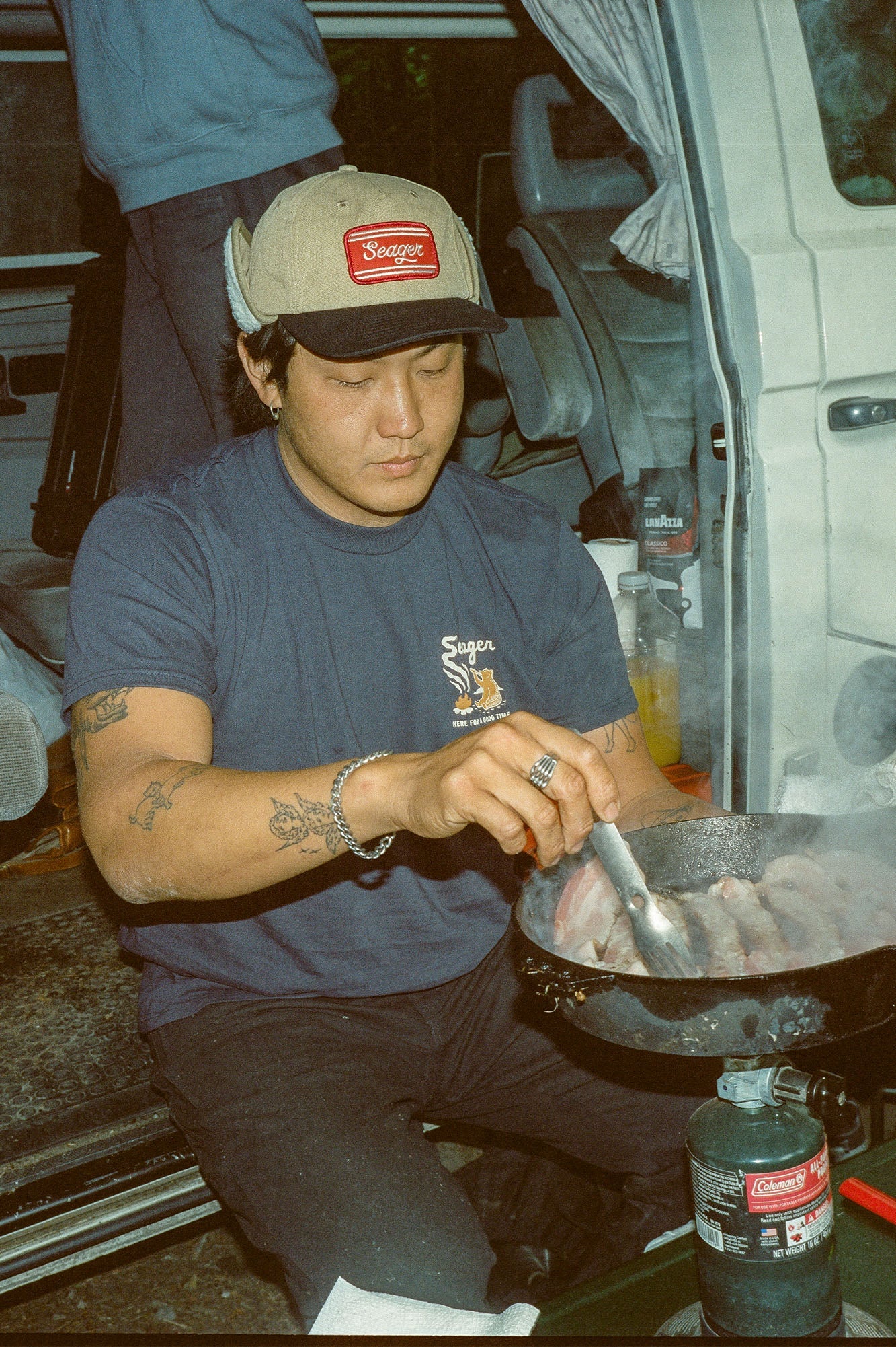 A man wearing the Smokey Tee in Navy by the brand Seager while he cooks out by a camper van