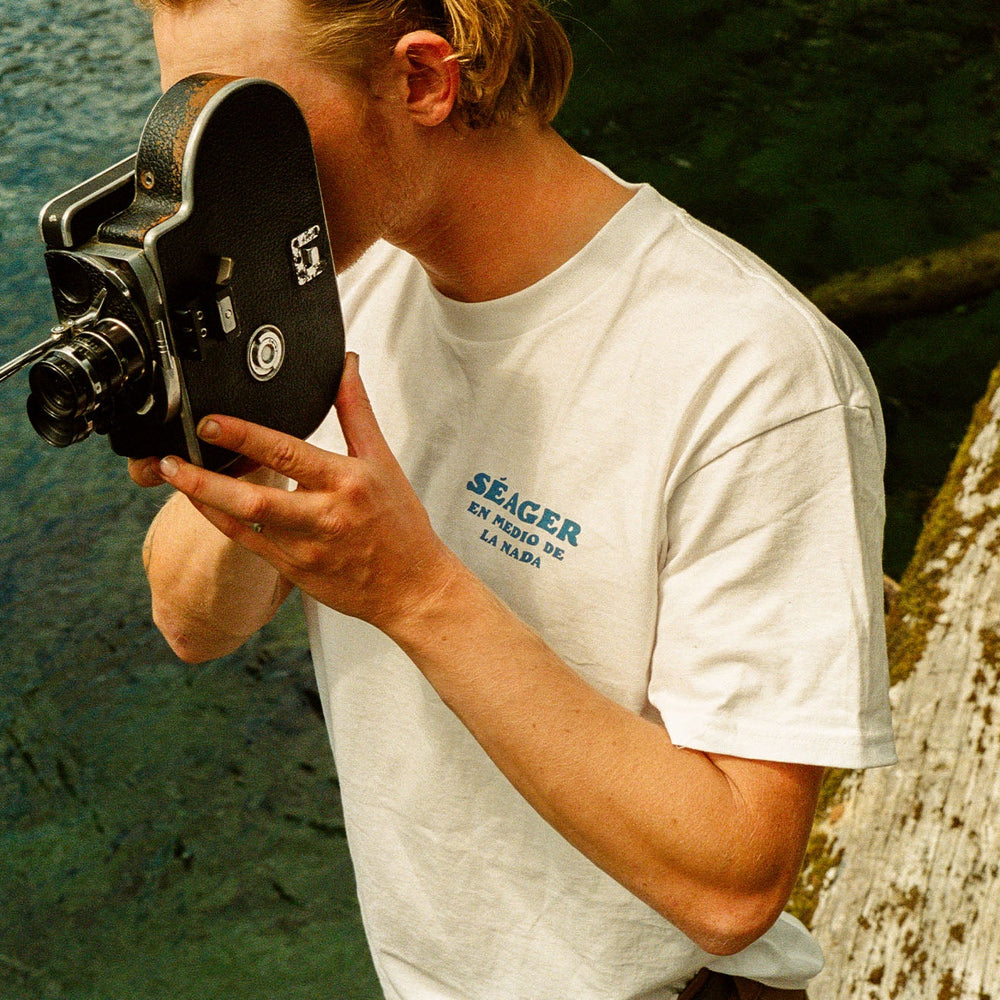 
                      
                        A man taking a photo of water on an old camera. He's wearing the white short sleeve Nowhere Tee from the brand Seager.
                      
                    