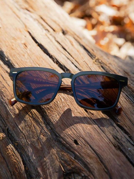 A pair of RAEN's Adin Men's Square Sunglasses with Cirus frames and Vibrant Brown Polarized lenses on a log