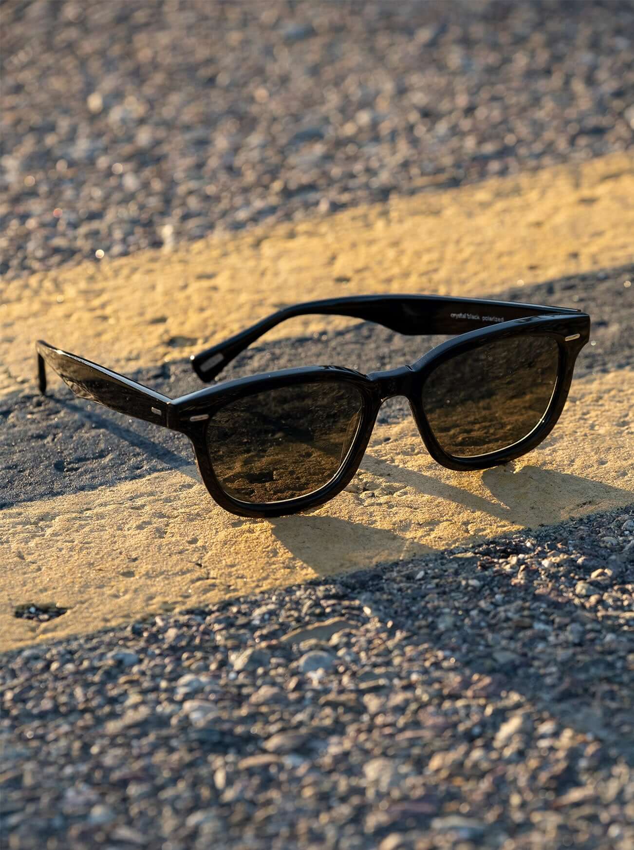 A pair of RAEN's Myles Square Sunglasses with Recycled Black frames and Green Polarized lenses on the street. 