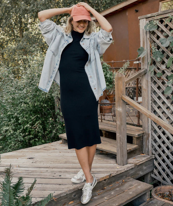 A woman wearing the Black Lexi Rib Mock Neck Midi Dress by Marine Layer. This dress offers versatile styling options and is paired here with a ball cap, sneakers and a denim overshirt