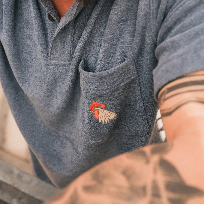 Close up front view of man wearing a plush blue short sleeve polo shirt with 2 button placket and single chest pocket with a rooster head design