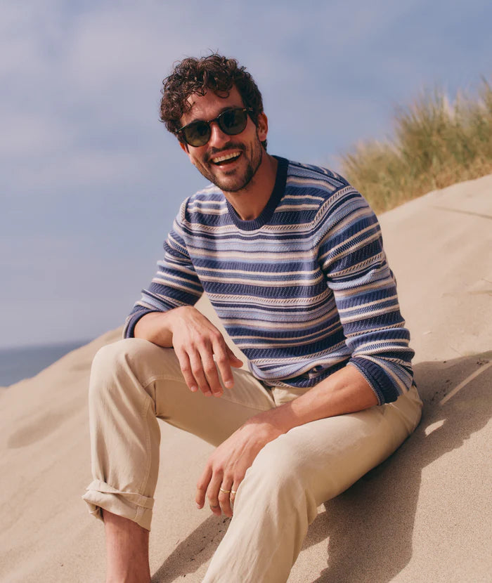 Man sitting on a sand dune while wearing the Blue Stripe Reed Jacquard Sweater by Marine Layer