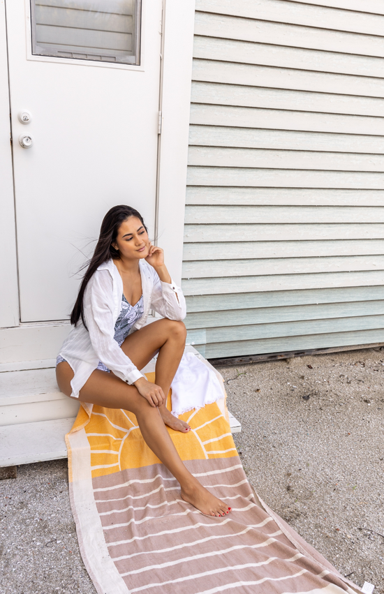 lifestyle photo of a model sitting on a sand cloud beach towel with a sun and sea inspired design and tassels at the ends