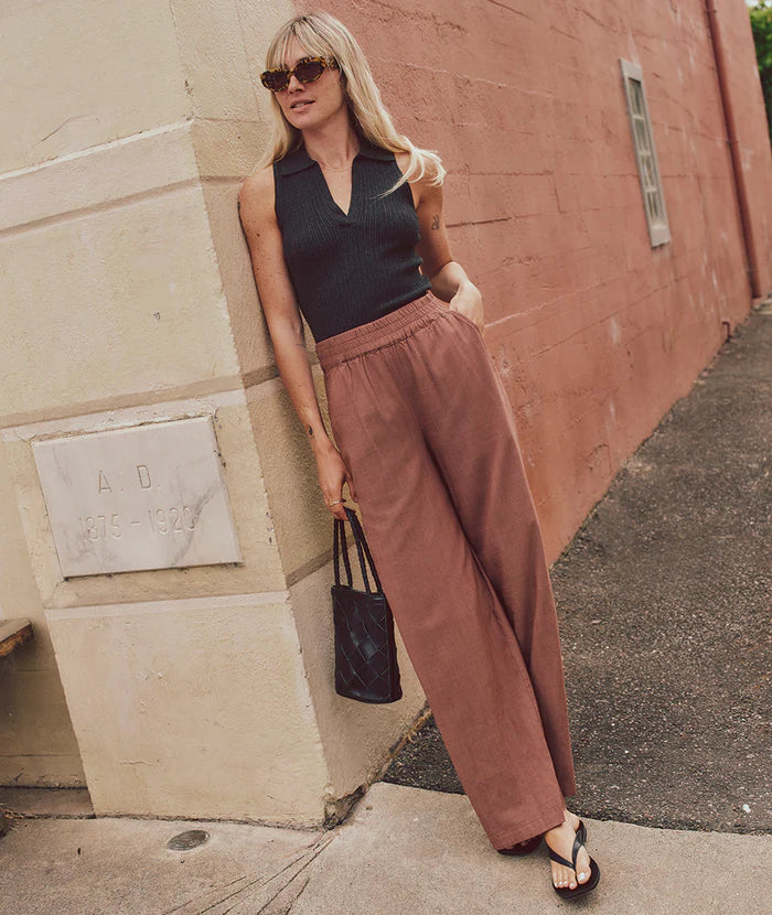 
                      
                        A woman leaning against a building is wearing the Pinecone color Allison Trousers by Marine Layer. They feature an elastic waistband and are paired here with a sleeveless black top and black flip flops. 
                      
                    