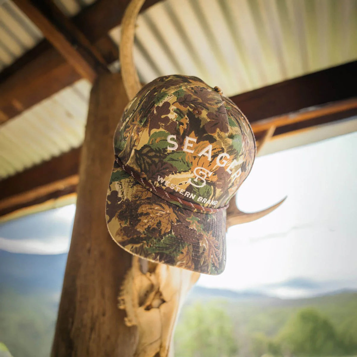 Camouflage cap with 'SEAGER' logo on a deer antler against a scenic background