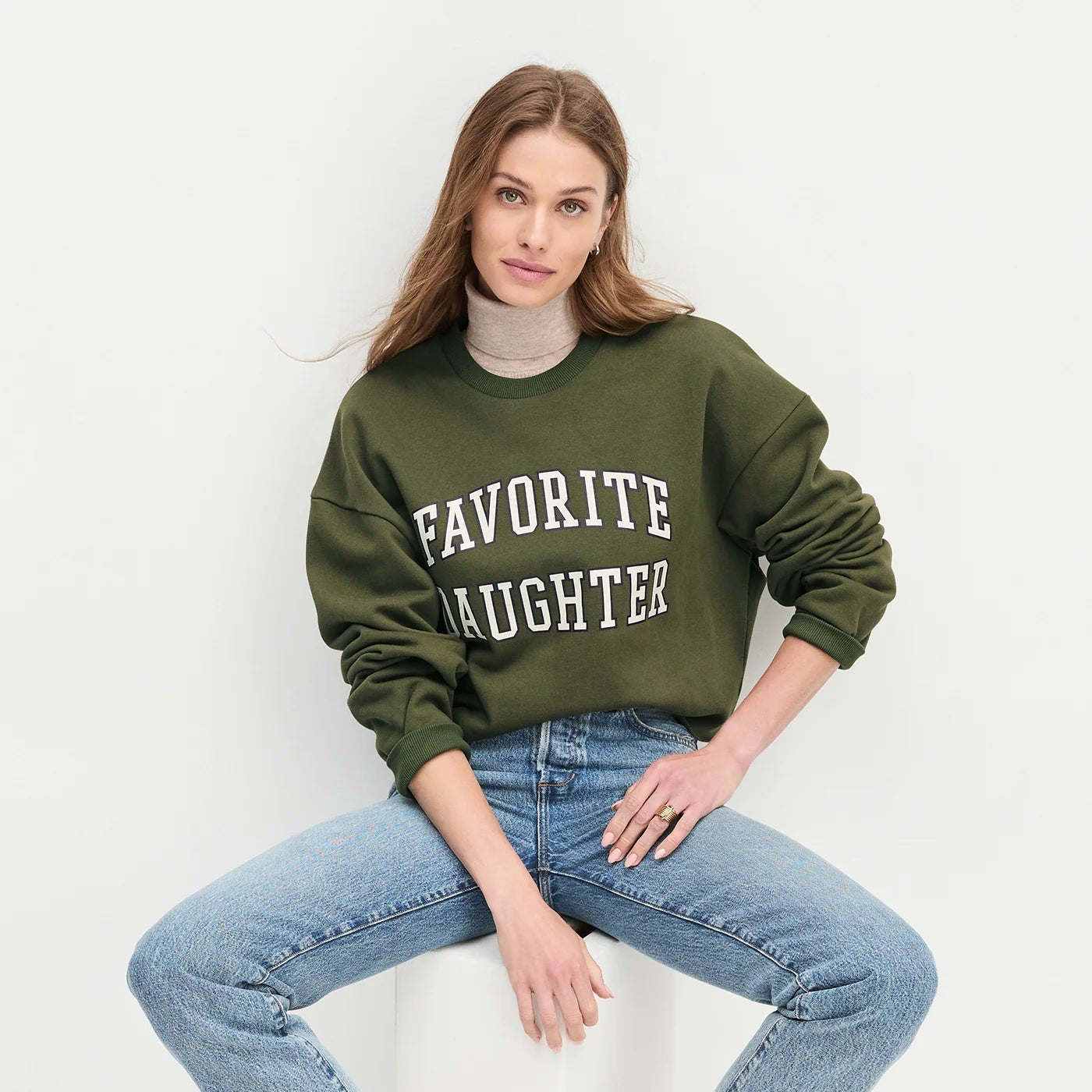 Woman wearing a green sweatshirt with 'FAVORITE DAUGHTER' text, sitting on a white stool 