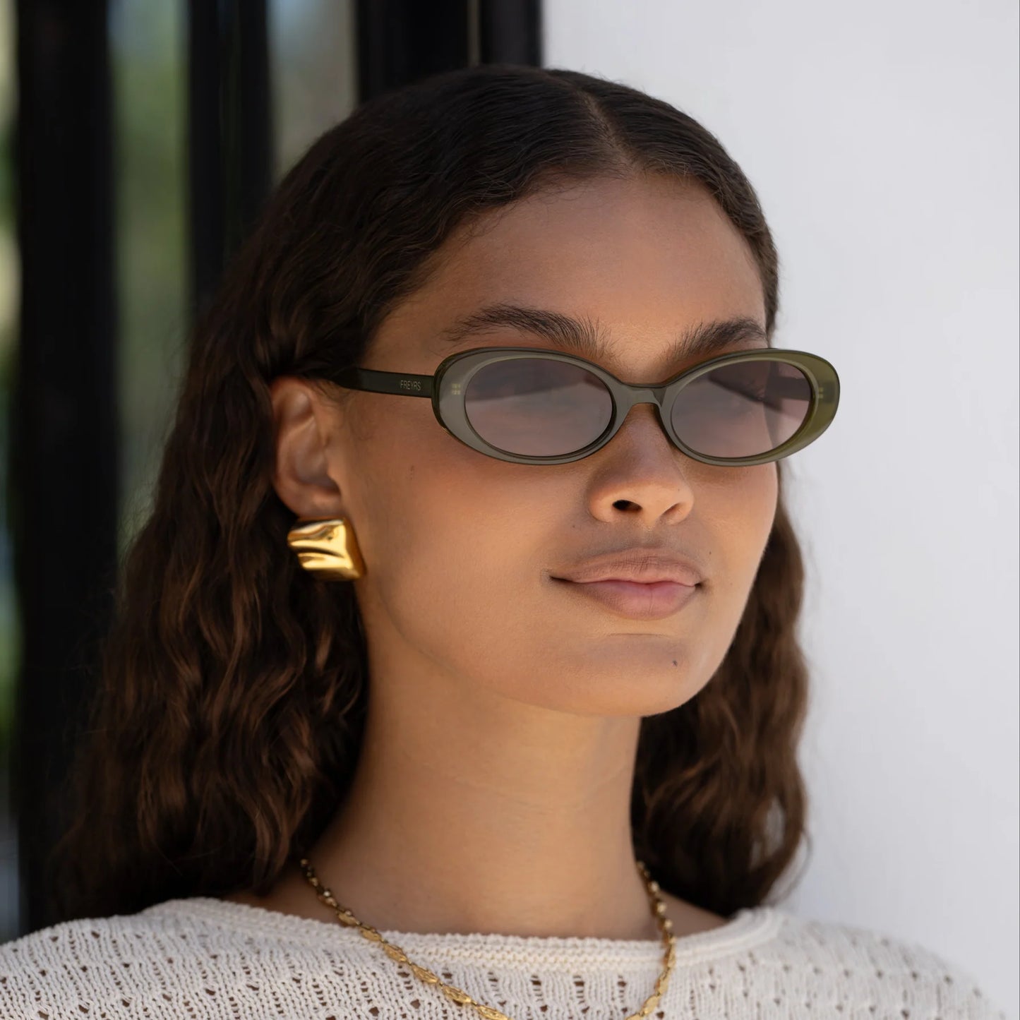 A woman wearing the FREYRS Mimi Women's Oval Sunglasses in the style Olive