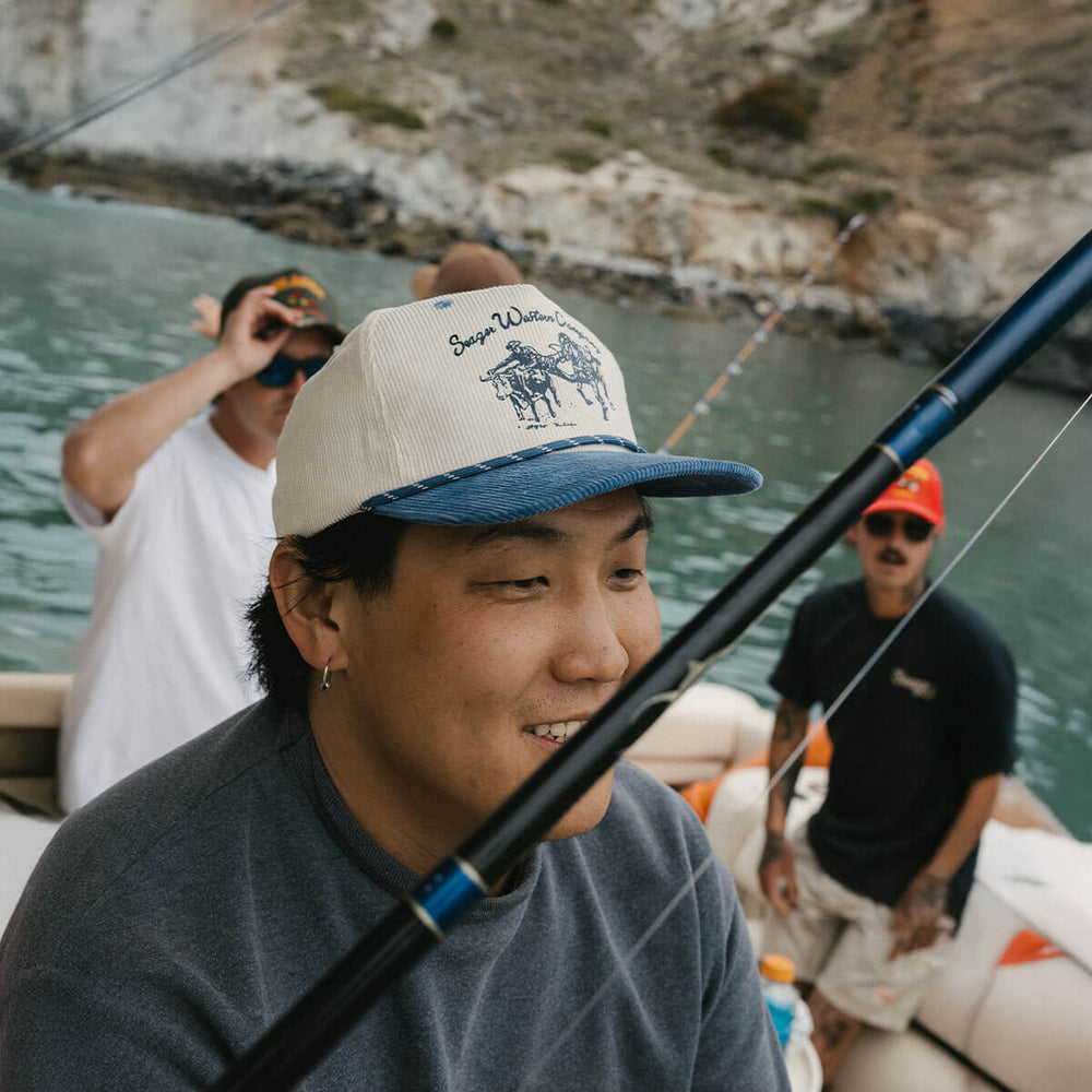 
                      
                        Man on a boat wearing a hat from Seager while holding a fishing rod with a scenic background
                      
                    