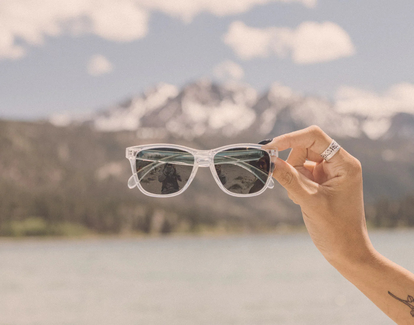 A person holding the Clear Forest Headland Sunglasses by Sunski in front of a mountain and lake